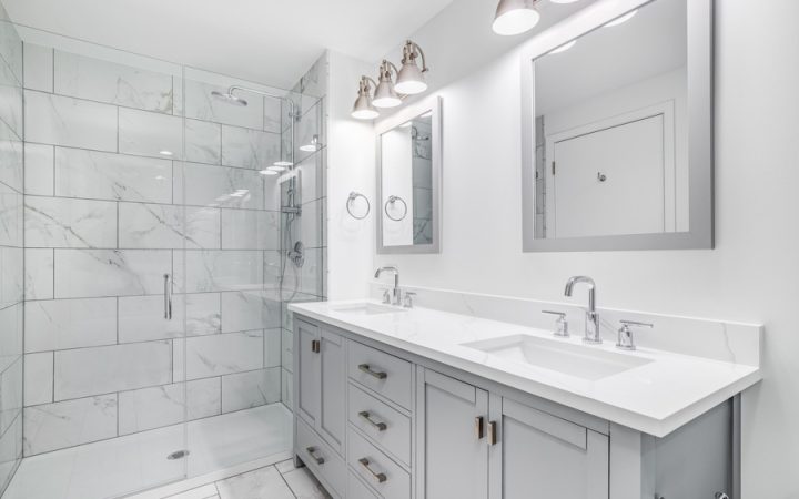 Modern bathroom designed with white marble tiles covering the floor and walls, featuring a dual sink vanity and a frameless glass shower, highlighted by stylish wall-mounted lamps.
