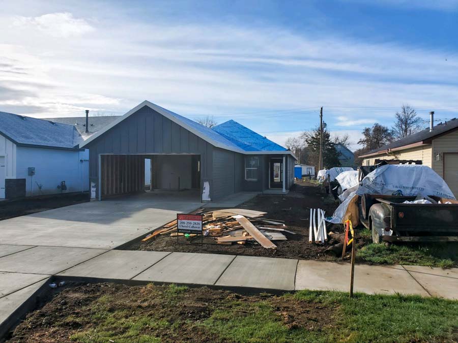 View of a residential home under construction with a freshly poured concrete driveway, blue tarps covering parts of the roof, and building materials scattered around.