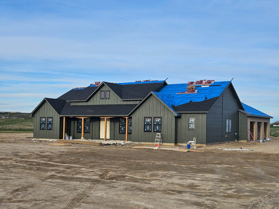 A large residential home under construction, painted dark green with a vivid blue protective roof covering, set against a rural landscape.