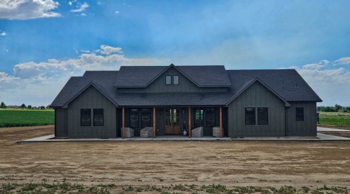 View of a newly completed residential home with gray siding, featuring a large driveway and set against a rural backdrop under a cloudy sky.