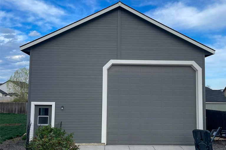 View of a newly completed residential home with gray siding, featuring a large driveway and set against a rural backdrop under a cloudy sky.