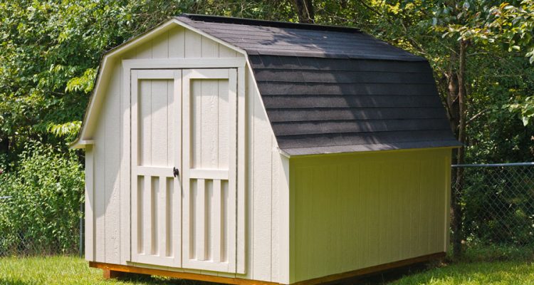 A small storage shed with a gambrel roof, situated on a grassy yard with trees in the background.